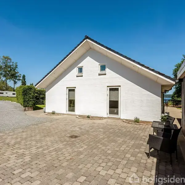 Hvidt hus med skråt tag står på en flisebelagt gårdsplads under en klar blå himmel. Omkring er der grusindkørsel og grønne buske samt træer i baggrunden.
