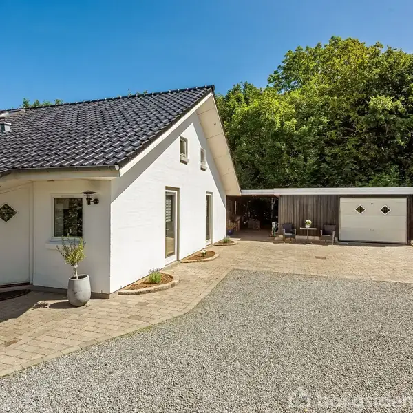 Hvidt hus med sort tag står langs en grusbelagt indkørsel. På den ene side er en carport og et skur. Omkringliggende grønne træer i baggrunden. Husnummer 52 ved døren.