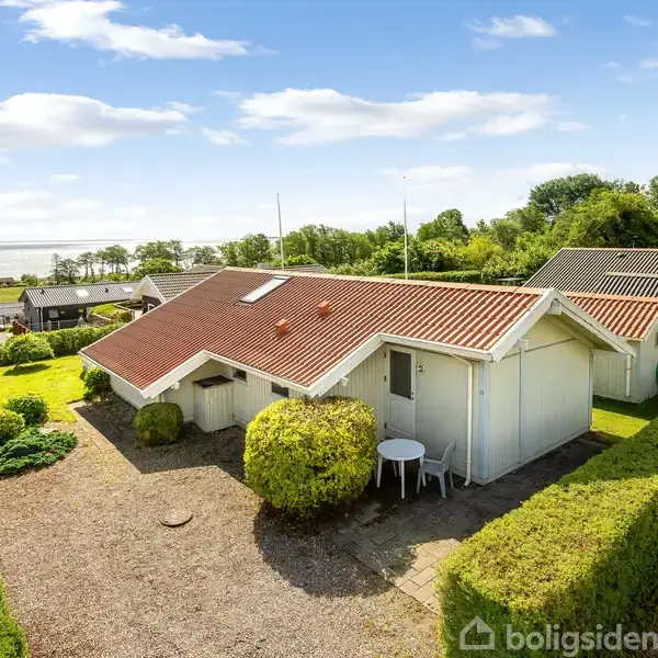 Et hvidt hus med rødt tag står omkranset af en veltrimmet hæk i en grøn have. I baggrunden ses flere huse og en udsigt til vandet.