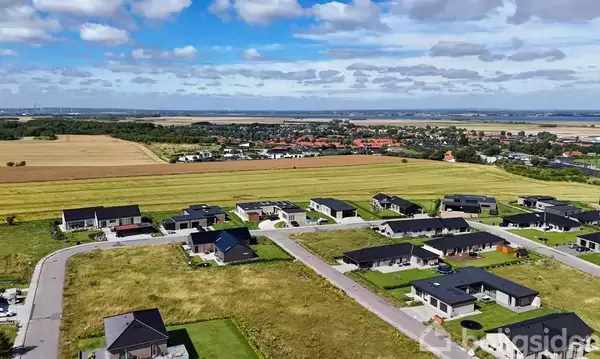 Boligkvarter med moderne huse ligger ved siden af åbne marker, under en delvist skyet himmel. I baggrunden ses en by og horisonten.