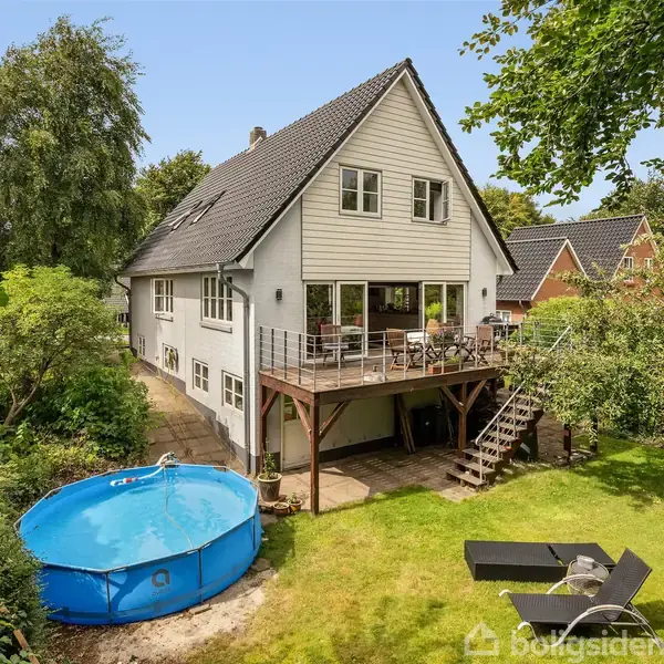 Et hvidt hus med skråt tag står omgivet af frodig have. Huset har en balkon, trappe og en pool i baghaven. Der er også liggestole på græsplænen.