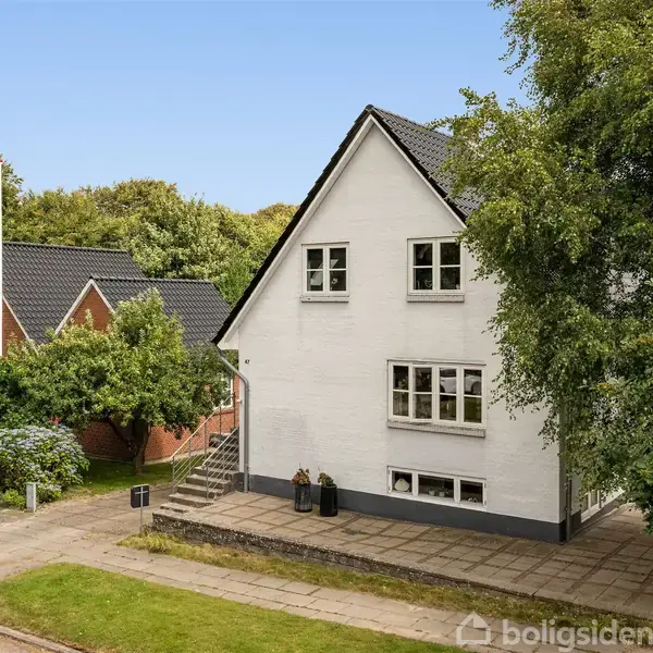 Hvidt hus med sort tag står på en træomkranset vej ved siden af et rødt murstenshus. Flisebelægning og trapper foran. Græsplæne og træer i forgrunden.
