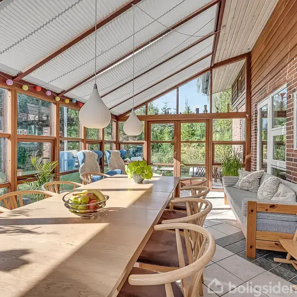 Et rummeligt udestueområde viser et langt spisebord i træ med stole, ophængte lamper, og udsigt til en frodig have gennem store vinduer. En sofa med puder står ved en murstensvæg.