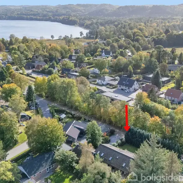 Et boligområde set fra oven med huse og træer i en frodig, grøn landskab. En rød pil peger mod et specifikt hus. I baggrunden ses en sø omgivet af skov.