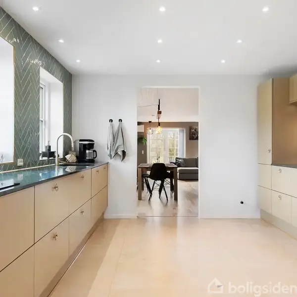Et moderne køkken med beige skabe, en sort bordplade og et grønt flisemønster som bagvæg. Køkkenet leder ind til en spisestue via en åben forbindelse. Lys indretning.