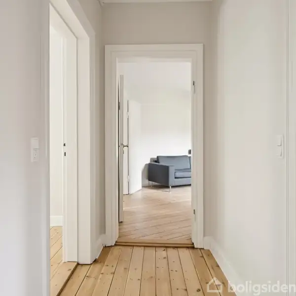 En gang med trægulv fører til et rum med en grå sofa. Hvide vægge og åbne døre skaber en lys og rummelig atmosfære.