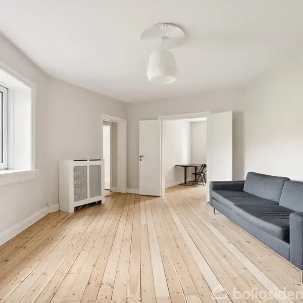 Sofa står i en lys stue med trægulv, ud mod vindue med hvid ramme. Baggrunden har åben dør til andet rum, hvor et lille bord ses.