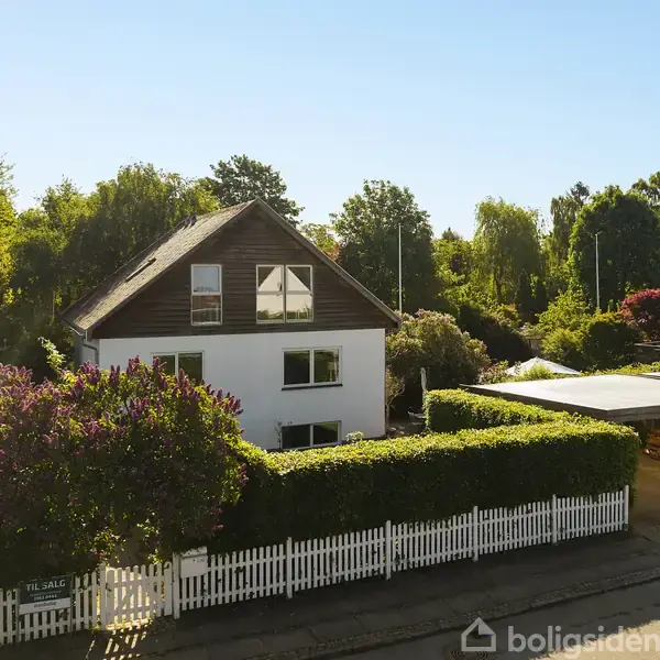 En hvid to-etagers villa med mørkt tag omgivet af frodig have og hvidt stakit. "Til salg" skilt i haven viser huset er på markedet. Solrig dag.