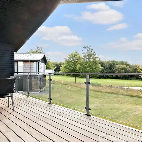 En træterrasse med glasrækværk har et havebord og stole. Terrassen har udsigt over en grøn græsplæne med træer i baggrunden under en blå himmel med skyer.