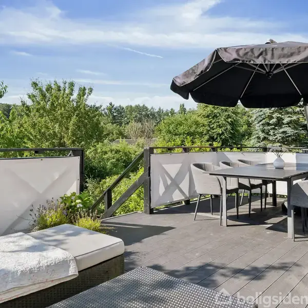 Terrassemøbler står under en stor sort parasol på en træterrasse, omgivet af frodig grøn beplantning og skovområder i baggrunden. En hvid polstret loungesofa er tæt på bordet.