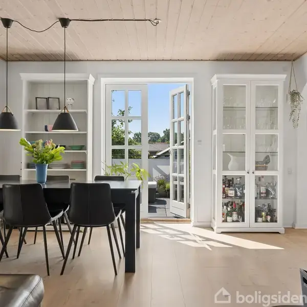 Spisestue med sort bord og seks stole, dekoreret med blomster i vase. Store vinduer og glasdøre til have. Hylder og vitrineskab med opbevaring langs væggene.