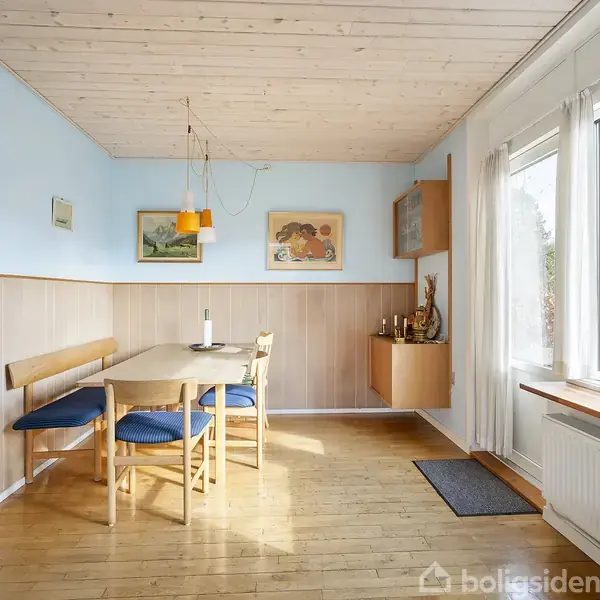 Stue med sofa, sofabord og stol på et tæppe foran store vinduer. Brændeovn ved væg udstråler hygge. Reoler fyldt med bøger og dekoration på væggen skaber en indbydende atmosfære.