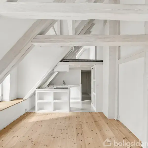 Et lyst rum med skrå lofter og synlige bjælker. Et lille, hvidt, åbent køkken står til venstre. Trægulv og vinduesnicher tilføjer charme. Stemningen er moderne og minimalistisk.
