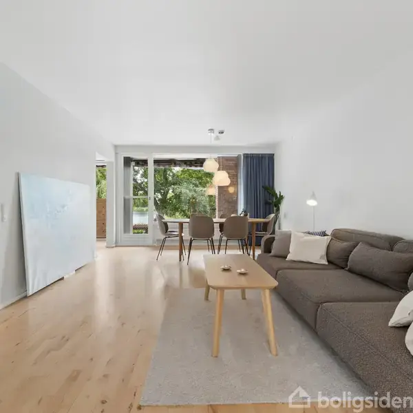 Gråt sofaarrangement med puder står i en stue, mens et træbord er centreret foran. Store vinduer i baggrunden giver udsigt til grønne omgivelser og en spiseplads.