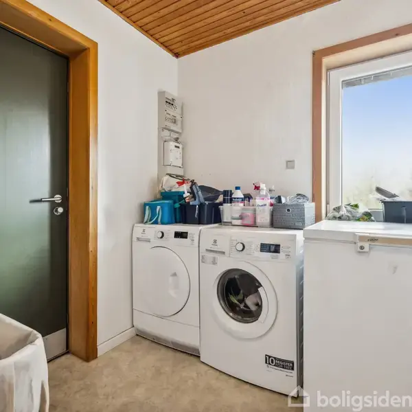 En vaskemaskine og tørretumbler står side om side i et lyst bryggers med træloft, ved siden af et vindue. Mange flasker og kurve er placeret ovenpå dem.