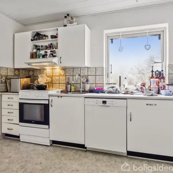 Køkken med hvide skabe og en ovn ved siden af en opvaskemaskine. Vinduet over bordpladen har hjerteformede dekorationer. Kaffemaskine og krydderier på bordet. Hundeskål på gulvet.