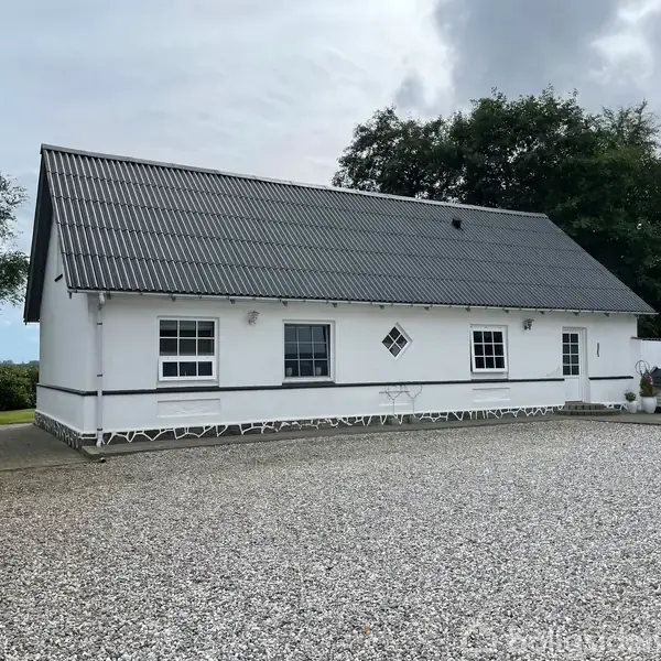 Hvidt hus står stille i en gårdsplads med grus omgivet af træer. Enkelt design med sort tag og små vinduer, der giver et rent og traditionelt udtryk.
