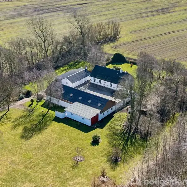 Gård med hvide bygninger og sorte tage omgivet af træer. Ligger på en stor græsplæne med marker i baggrunden.