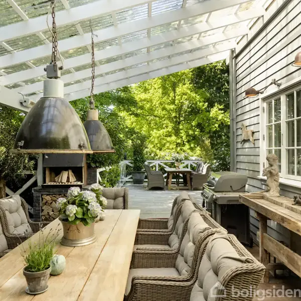 Et overdækket udendørs spiseområde med en lang træbænk og polstrede stole. To store lamper hænger over bordet. I baggrunden er en grøn have.
