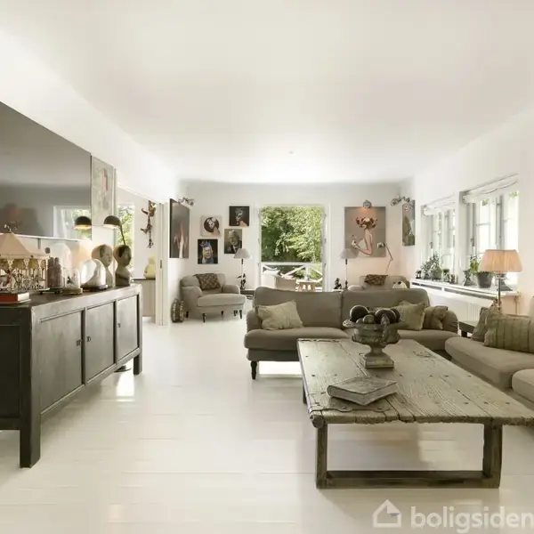 En lys stue med en grå sofa og træbord, dekoreret med kunstværker på væggene. Store vinduer lader naturligt lys strømme ind, og grønne planter skaber en hyggelig atmosfære.