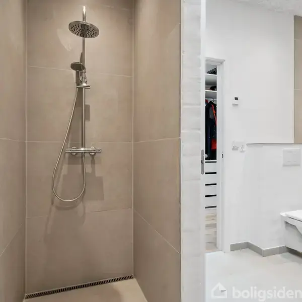 Bruseniche med moderne bruser hænger over beige fliser; ved siden af er et lyst badeværelse med væghængt toilet og åbning til et walk-in closet.