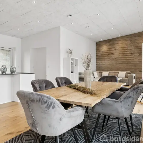 Et spisebord med grå polstrede stole står i et moderne, lyst køkken-alrum. Rummet har trægulve, en sofa i baggrunden, og en væg med træpaneler til højre.