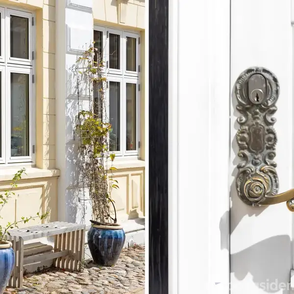 Dør med udsmykket håndtag står åben; udsigt til en gårdsplads med brosten, to blå keramikkrukker med planter og en gammel gul murstensbygning med hvide vinduer i baggrunden.