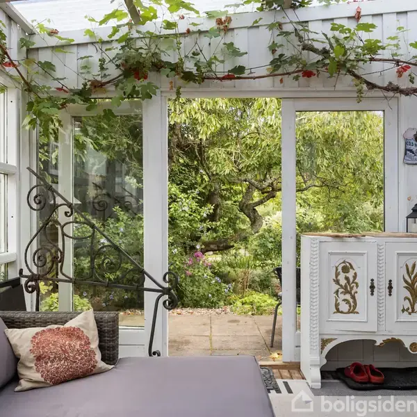 En hvid udestue med glasvægge har en sofa med puder til venstre og et dekoreret skab til højre. Baggrunden viser en frodig have med grønne planter.