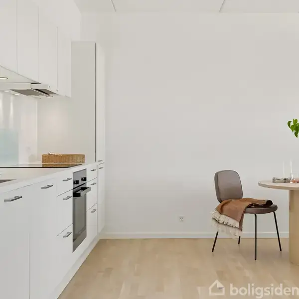 Et hvidt, moderne køkken med integrerede skabe og ovn, står ved siden af et rundt træbord med en stol. Der står en vase med grønne planter på bordet.
