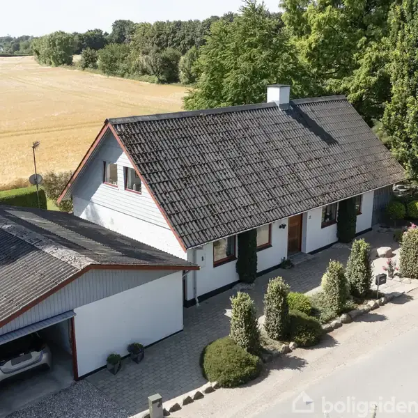Et hvidt hus med sort tag står ved siden af en garage, omgivet af hække og træer. I baggrunden ses en gylden mark og en skov.