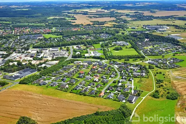 En række boliger ses fra luften, omgivet af grønne marker og skove. Bygninger er arrangeret i et mønster med veje imellem, og en by ligger i baggrunden.