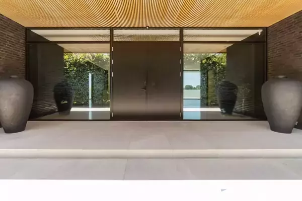 Døråbning udført i mørkt glas med to store keramikkrukker ved siden af. Omkringliggende flisebelægning og murstensvægge. Refleksion af grønne planter udenfor.