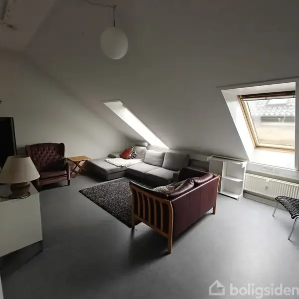 En stue med en grå hjørnesofa og et brunt læderstole står under et skråvindue. Rummet er lyst, med gråt gulv, en gulvlampe og en hvid radiator.