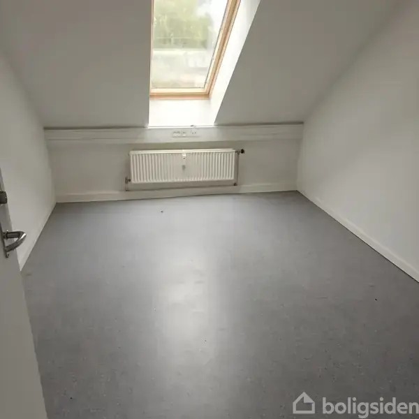 Et tomt rum med et skråt loft og et ovenlysvindue. Der er en radiator under vinduet. Gulvet er dækket af grå linoleum, og væggene er hvide.