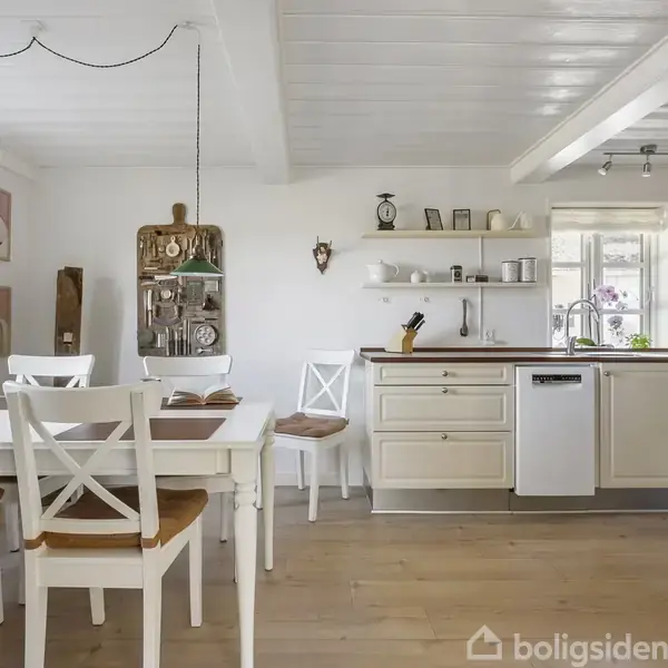 Et lyst, rummeligt køkkenalrum med hvidt bord og fire stole til venstre. Køkkenet til højre har hvide skabe og en opvaskemaskine. Rummet har trælofter og -gulve. Ingen synlig tekst.