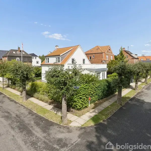 Hvidt hus med rødt tag står på hjørnegrund omkranset af hæk og træer, placeret i et villakvarter med klare blå himmel og flere nabohuse i baggrunden.