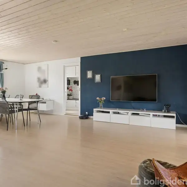 Stue med fladskærms-TV på mørk blå væg, hvide møbler, spisebord med stole til venstre, store vinduer, lyse trægulve og hyggebelysning.