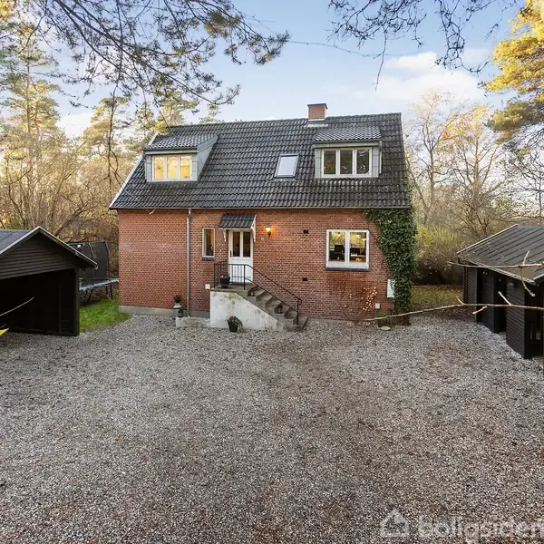 Et rødt murstenshus står stille omgivet af grus og høje træer. To sorte garager flankerer indkørslen, der leder op til trappen ved hoveddøren.