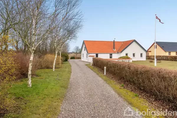 Hus med rødt tag står fredeligt i landskab. Grusvej fører dertil, omgivet af buske og græs. I baggrunden ses en flagstang med Dannebrog og flere træer.