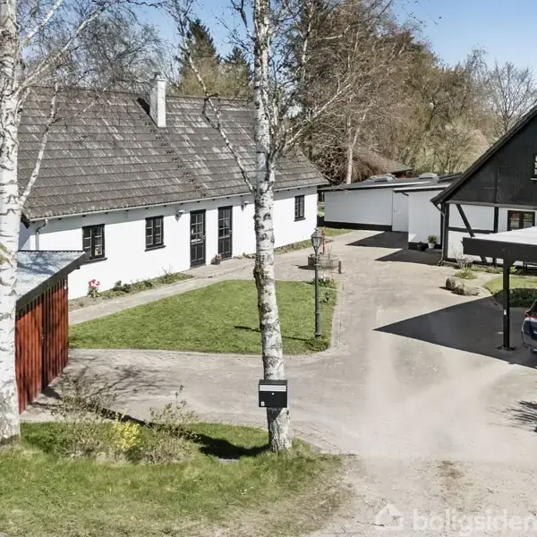 Hvidt hus med mørkt tag står stille ved grusvej omgivet af træer og en carport med en bil. Grønt græsareal og rød tilbygning ses ved siden af.