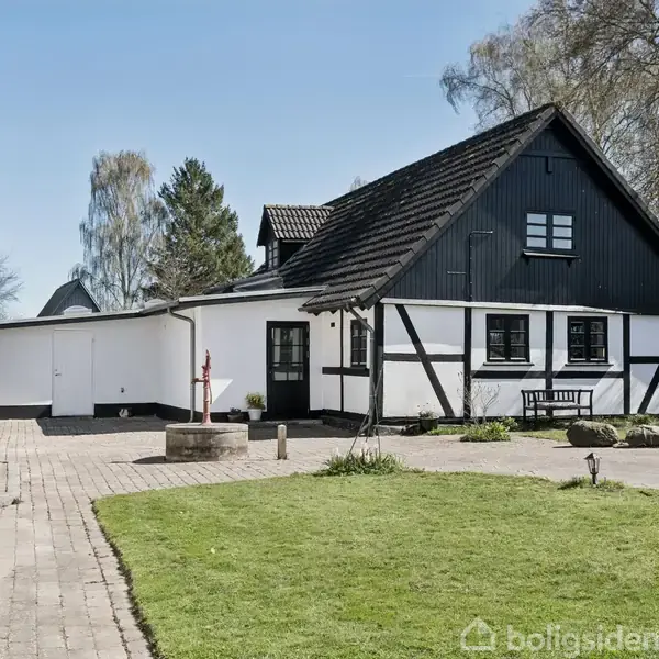 Et hvidt bindingsværkshus med sort tag står stille i en have med græsplæne og flisebelagt sti. Træer omgiver ejendommen under en klar blå himmel.