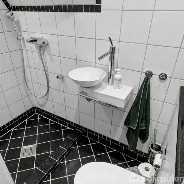 Brusebad og vask i et badeværelse med hvide fliser på væggene og sorte fliser på gulvet. Der hænger et grønne håndklæde ved vasken, og der er en toiletpapirholder.