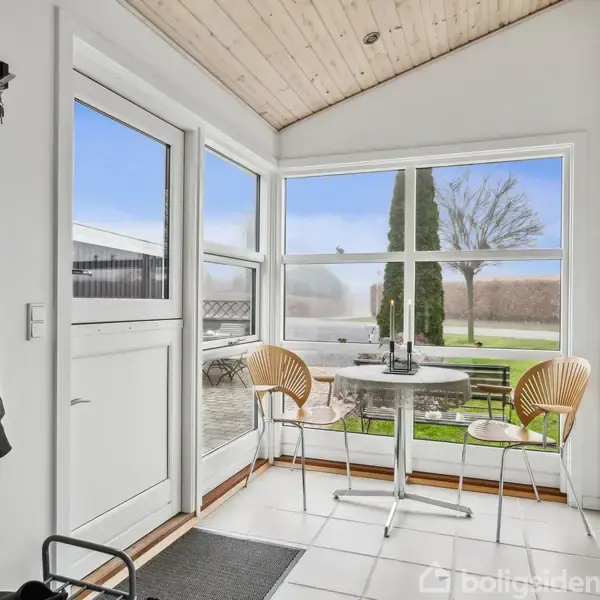 Et lille rundt bord med to stole står i et lyst rum med store vinduer, der viser en have med træer udenfor. Jakker hænger ved døren på en knagerække.