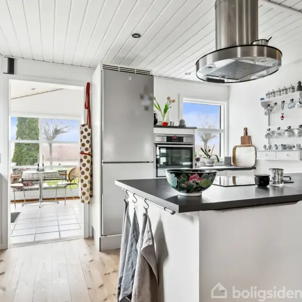 Et moderne køkken med hvid bordplade og køkkenø har en stor emhætte. Rummet til højre viser åbne hylder og en indbygningsovn. Udsigt til stue med lysindfald.