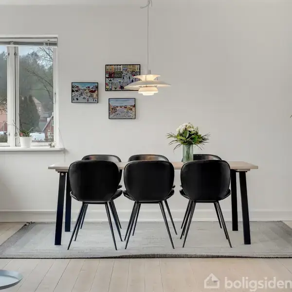 Et spisebord med seks sorte stole står på et tæppe. En vase med blomster er midt på bordet. Væggene er dekorative med billeder, og et vindue viser en udendørs udsigt.