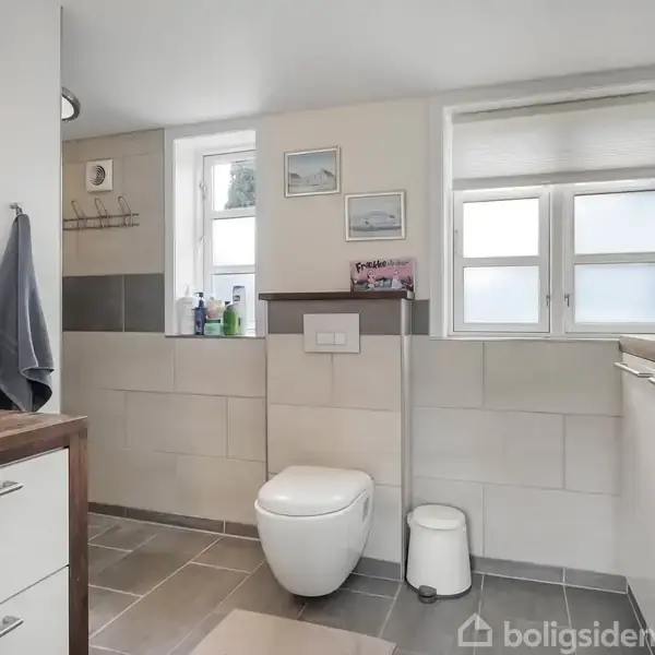 Toilet med skjult cisterne under vindue nær brusesektion; håndvask og skab til venstre; gråt flisegulv og hvidt vægflise design, hygiejneartikler placeret på hylder.