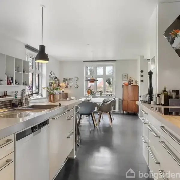 Køkken med hvide skabe og stålbordplader, to hængende lamper over bordpladen, åbner op mod spisestue med gråt spisebord, moderne stole, træskab, og vinduer med udsigt til træer.