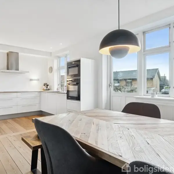 Et lyst køkken med et træbord og sorte stole i forgrunden. Rummet har lyse trægulve, hvide skabe og store vinduer mod en udsigt over hustage.