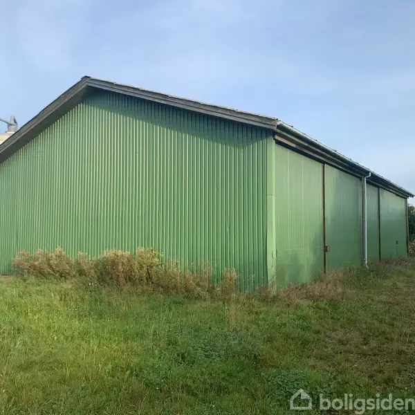 En stor, grøn lagerbygning står stille i et landligt område, omgivet af græs og buske. Skylinen er domineret af en lyseblå himmel.