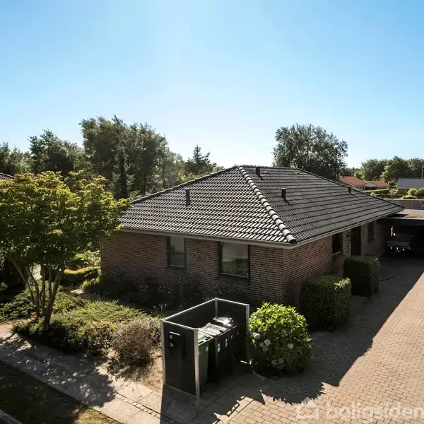 En etplans murstenshus står stille i et solrigt boligkvarter. Forhaven er velplejet med træer og buske, mens en flisebelagt indkørsel fører til en carport til højre.